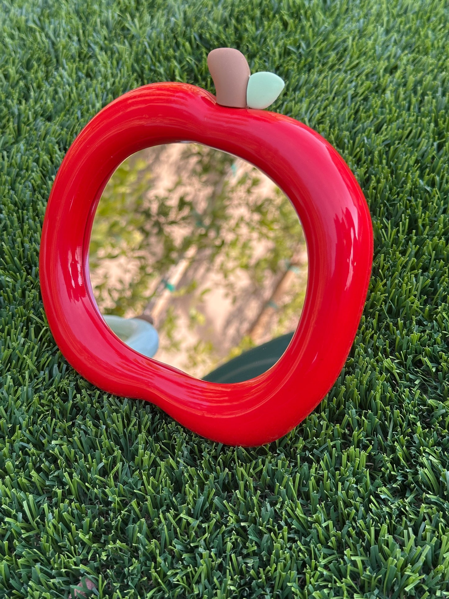Two apple-shaped makeup mirrors, one red and one green, wall-mounted, placed on grass with a blurred background.